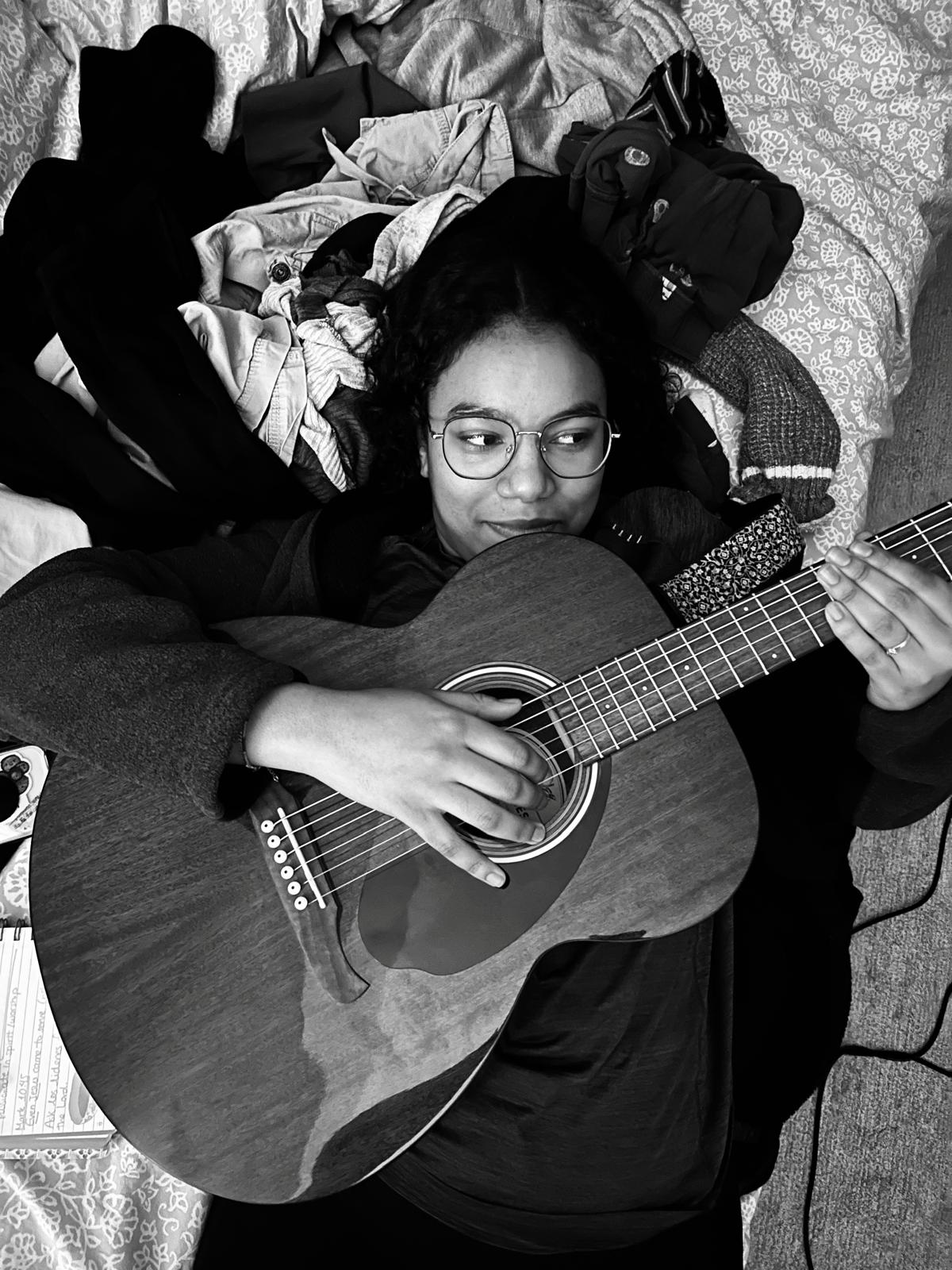Mia lying on her bed playing acoustic guitar, songwriting notebook beside her, black and white